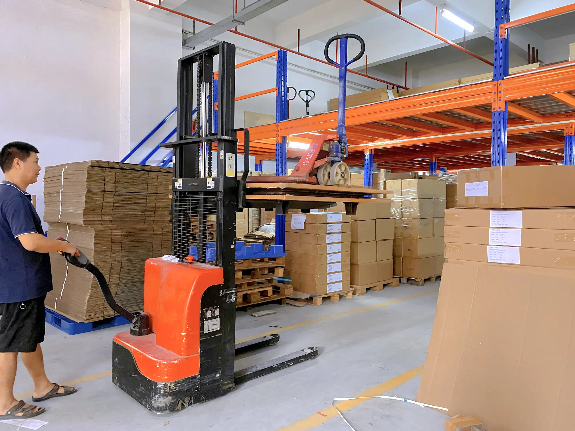 Corrugated board rolls and stacked board sheets inside a display factory warehouse, with different flute grades labeled and a forklift moving a pallet of double wall board, showing the raw material stage before display production begins.