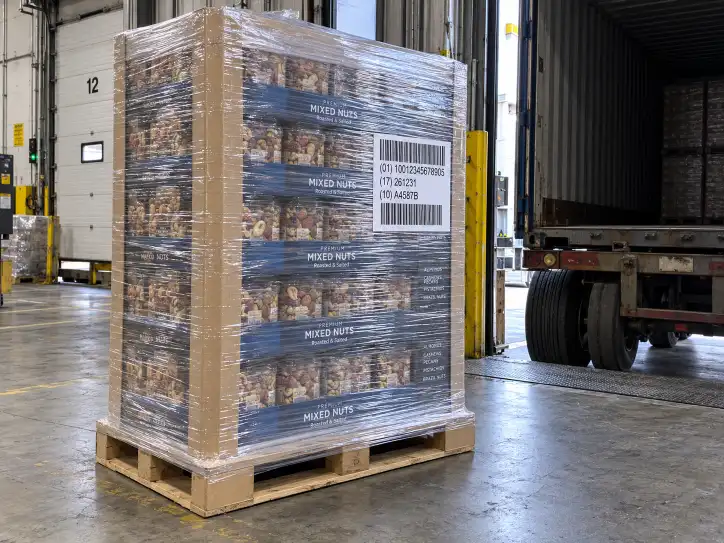 Pre-filled Costco pallet display wrapped in stretch film with corner boards on all four vertical edges and a GS1-128 pallet label visible on the side, standing in a factory shipping bay beside a container trailer, showing the transit-protected final unit.